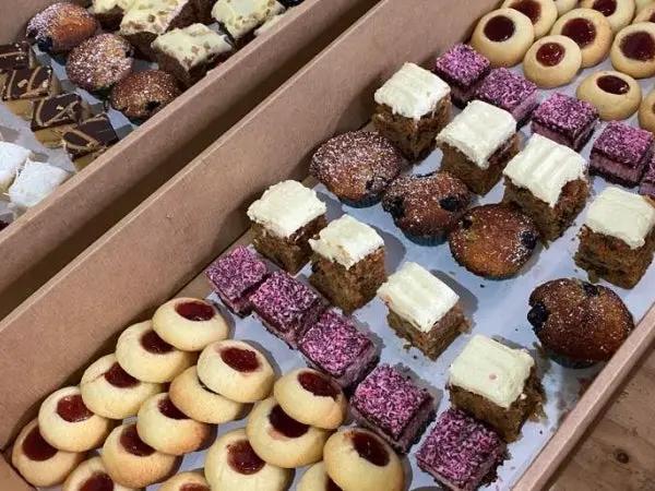 A close-up view of assorted baked goods arranged in cardboard trays, including round jam-topped cookies, square frosted cakes, and chocolate-dusted pastries on a light surface.
