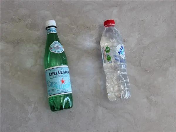 Two plastic water bottles lying on a gray stone surface: a green glass bottle of San Pellegrino with a white cap and light blue label, and a clear plastic bottle of Aquafina with a red cap and green label.