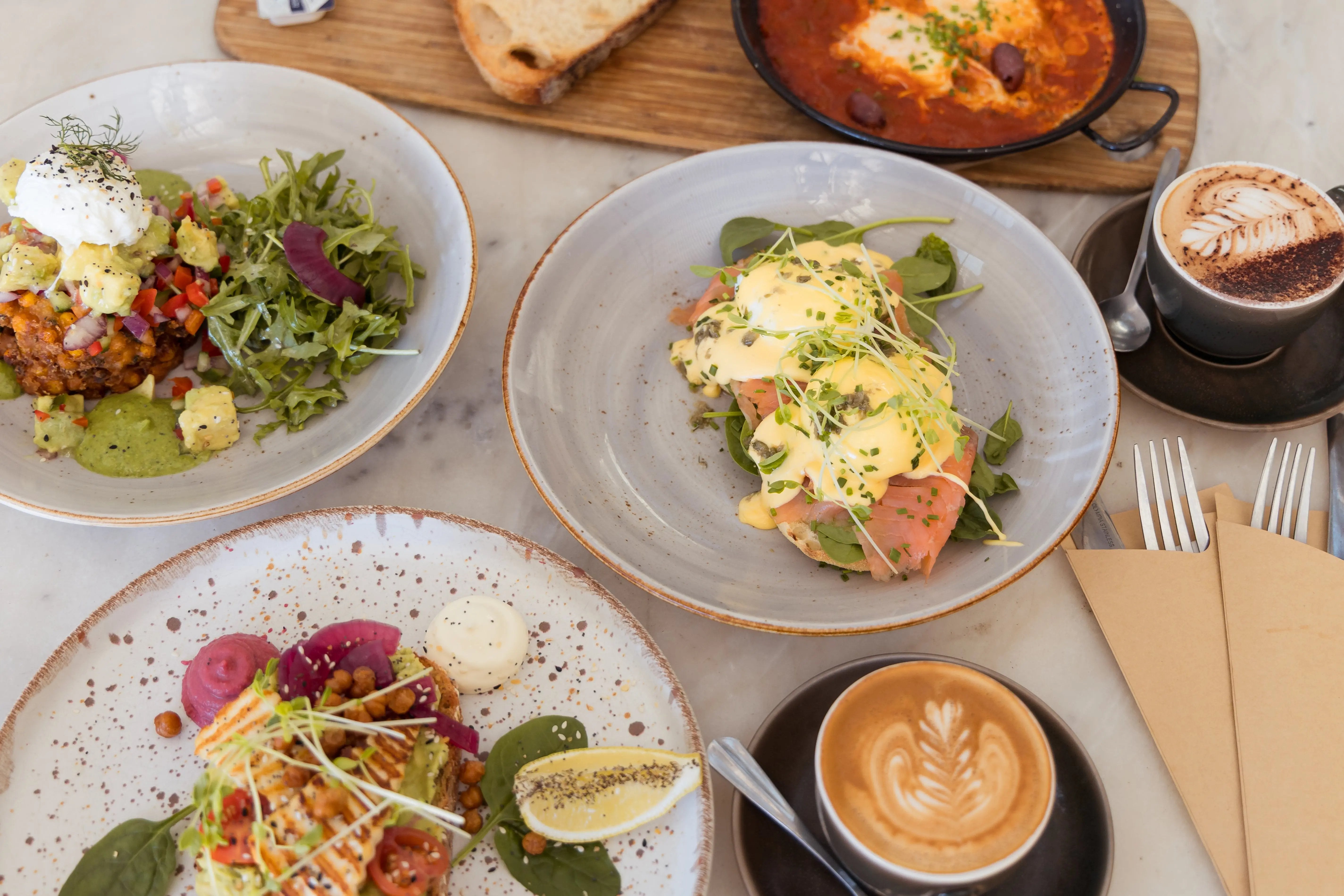 A top-down view of a breakfast table setting featuring multiple dishes: a plate of eggs with hollandaise sauce, smoked salmon, and microgreens; another plate with a salad and fried item with sour cream; a cup of latte with leaf art; and a cup of coffee with cocoa powder on a marble tabletop.