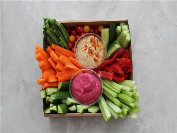 A top-down view of a square wooden serving tray filled with colorful vegetable sticks and two small bowls of dip on a light gray surface. The tray contains carrot sticks, cucumber sticks, red pepper strips, asparagus, cherry tomatoes, and two dips: one beige with red seasoning and one bright pink.