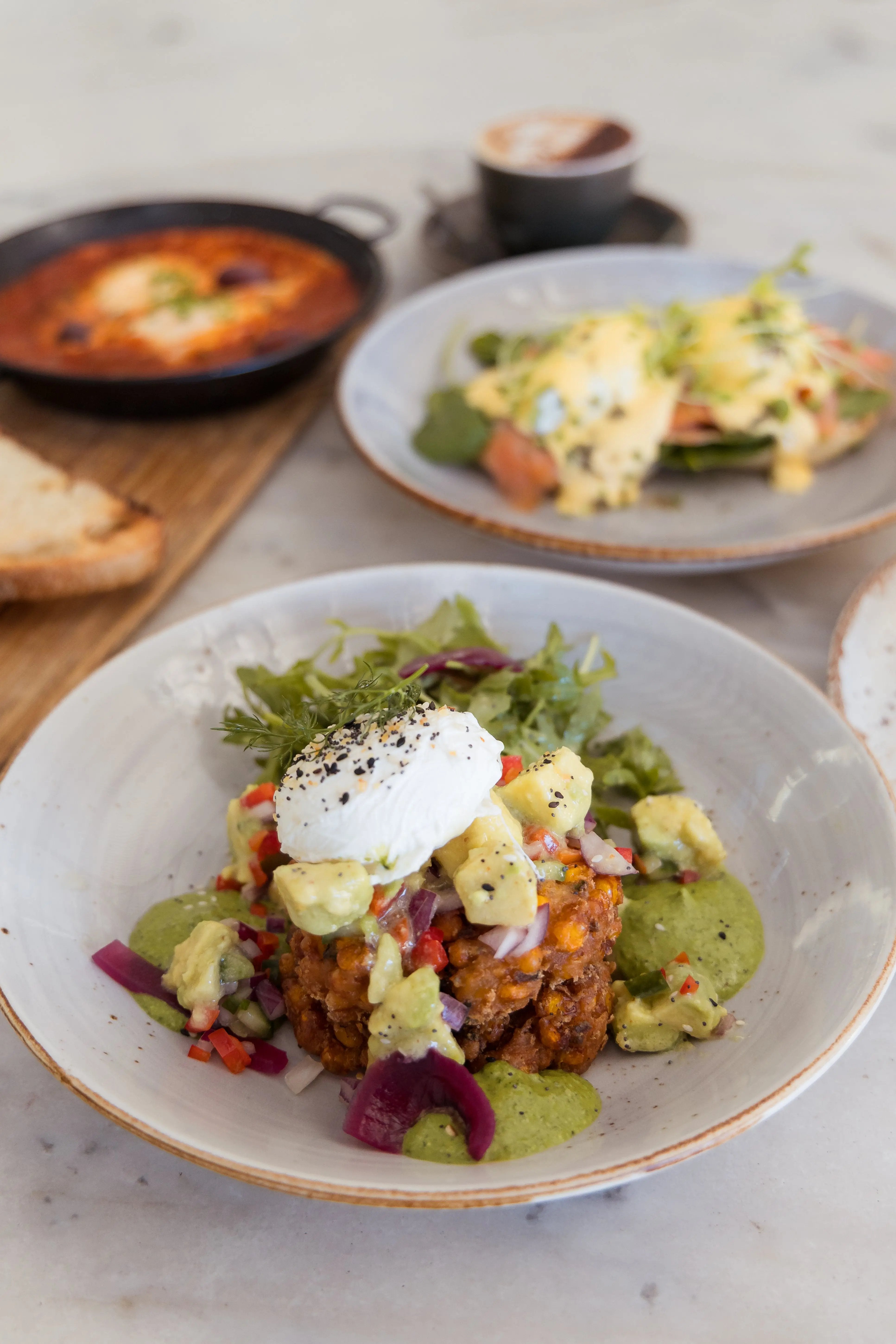 A plated breakfast dish featuring corn fritters topped with a poached egg, avocado, red onion, and green sauce, served on a light ceramic plate. Another plate with a yellow omelet and a black skillet with an orange stew are blurred in the background on a light marble surface.