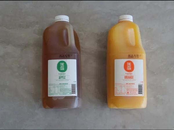 Two large plastic jugs of juice from The Farm, one apple juice in a bottle and one orange juice in an orange-yellow bottle, placed side by side on a gray concrete table