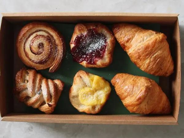 A top-down view of a cardboard box containing six assorted pastries, including two croissants, a cinnamon swirl, a jam-filled pastry, and a lemon tart, arranged on a dark green liner over a light marble surface.