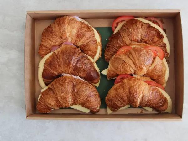 Six croissant sandwiches filled with cheese and tomato slices, arranged neatly inside a brown cardboard box on a light gray surface.