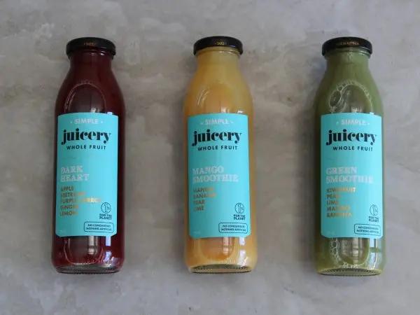 Three glass bottles of juice from the brand 'juicery whole fruit' lined up on a light gray stone surface. The bottles contain dark red, yellow-orange, and green juices, each with a light blue label and a black cap.