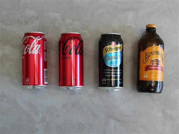 Four beverage containers arranged in a row on a light gray stone surface: two red Coca-Cola cans, one black Schweppes Lemonade can, and one brown glass bottle with an orange label reading Bundaberg Ginger Ale.