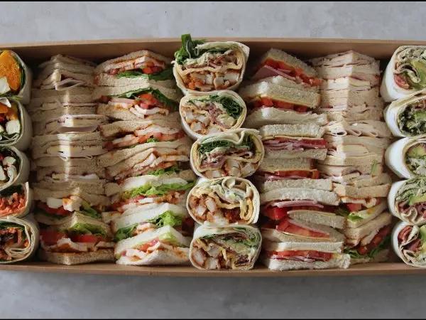 A large cardboard tray filled with assorted prepared sandwiches and wraps, including club sandwiches, turkey wraps, and tomato and cheese slices, photographed from above on a light surface.