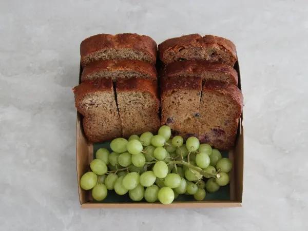 Two rectangular boxes of sliced bread or cake, each containing several thick slices stacked vertically and a bunch of green grapes, set on a light gray marble-like surface.