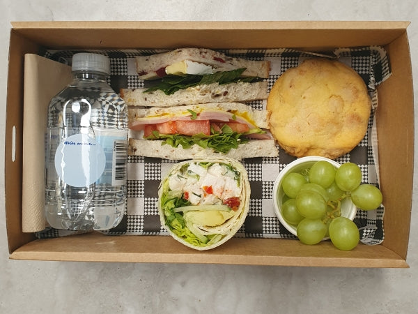 A lunch hamper box containing a gourmet wrap, a gourmet sandwich cut into triangles, a house made cookie, seasonal fruit (grapes), a 250 ml water bottle, and a napkin.