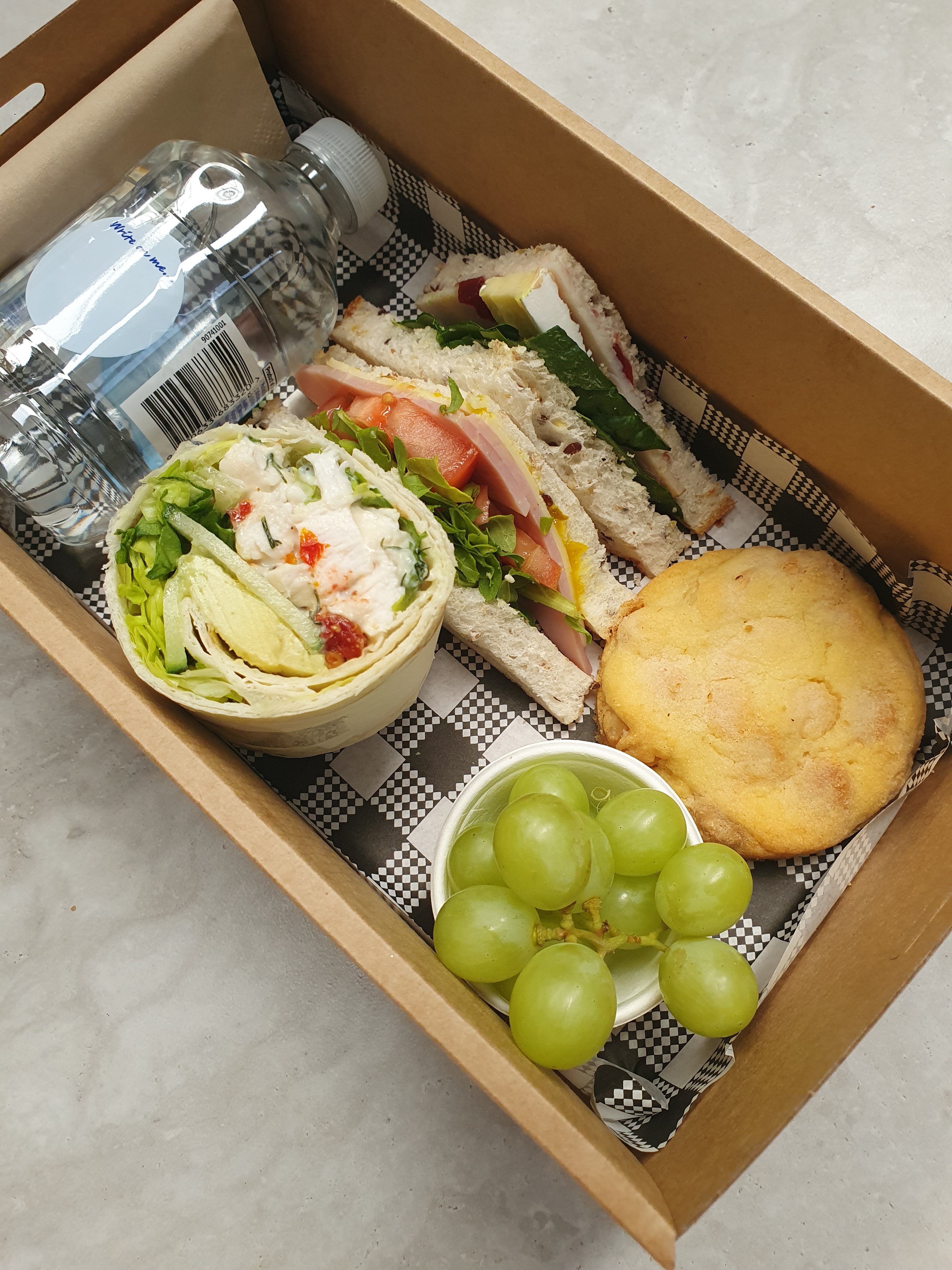 A lunch hamper box containing a gourmet wrap, a gourmet sandwich cut into triangles, a house made cookie, seasonal fruit (grapes), a 250 ml water bottle, and a napkin.