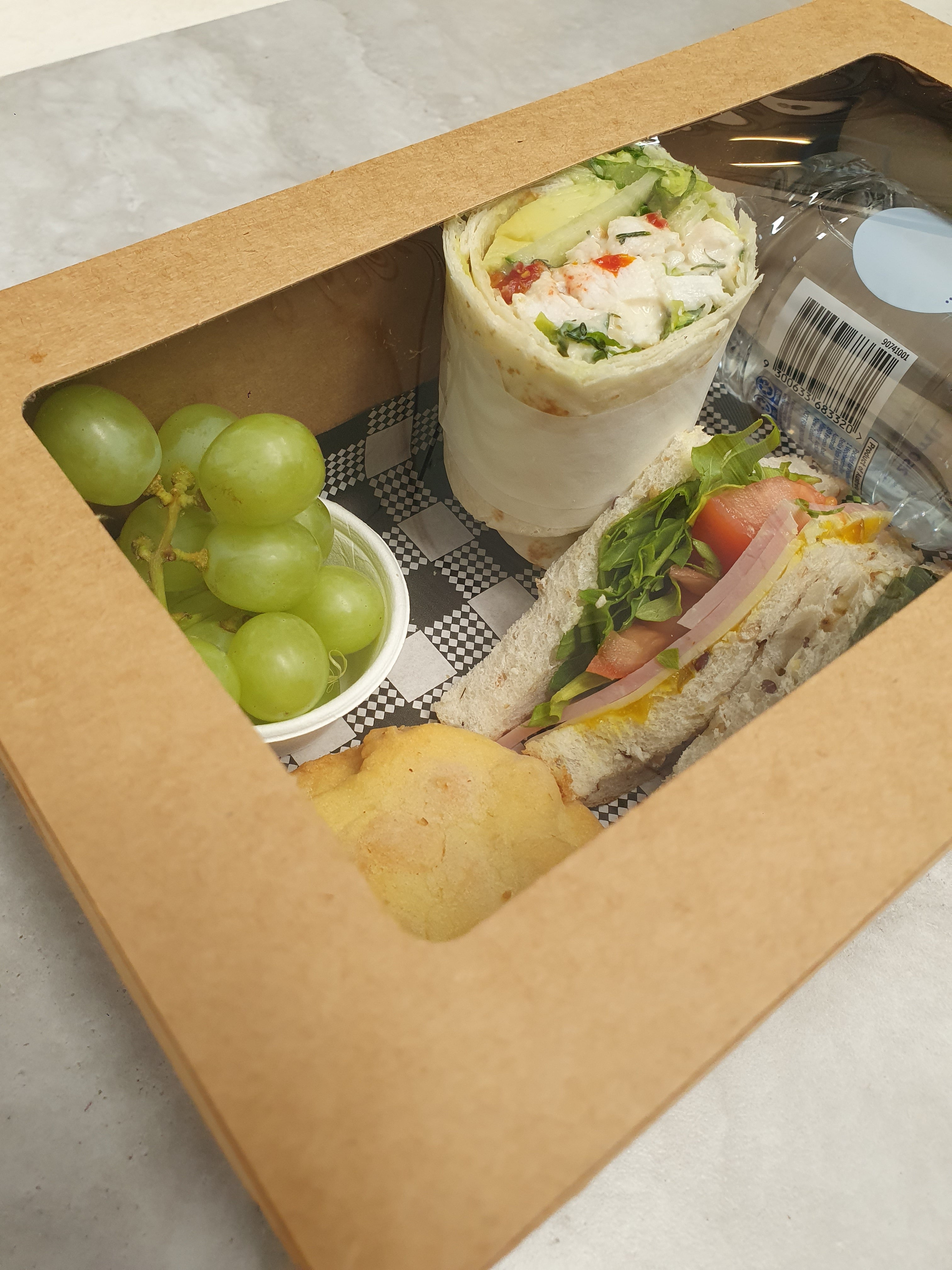 A lunch hamper box containing a gourmet wrap, a gourmet sandwich cut into triangles, a house made cookie, seasonal fruit (grapes), a 250 ml water bottle, and a napkin.